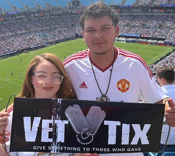 woman and man holding Vet Tix sign in a soccer stadium. Give some thing to those who gave